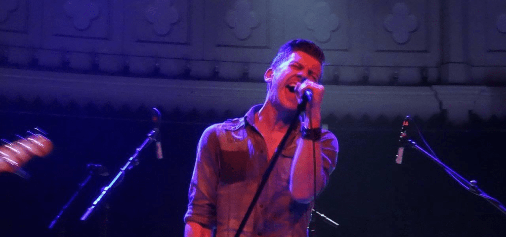 A white man with spiky hair, standing on stage bathed in pink-purple light. He's singing (clearly with passion) into a microphone.