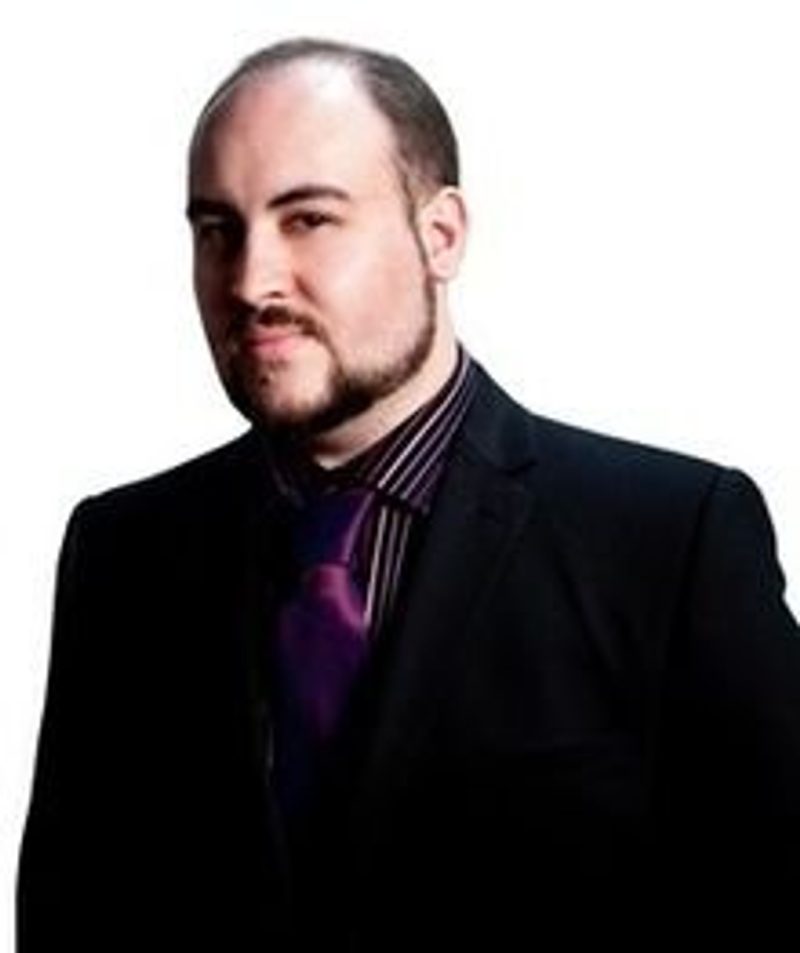 A white man in his 30s, with a neatly trimmed narrow beard and nearly shaved head is wearing a black suit with a purple button-up shirt under it. He's half smiling.