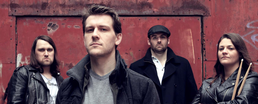 3 white men and one white woman standing in front of a rusty old wall. The first man has long hair and is wearing a leather jacket. The second is further forward in a grey coat. The third man is in a peaked cap and a black peacoat. The woman is in a leather jacket, holding drumsticks. They're all dressed for UK autumn weather, looking all mean and moody.