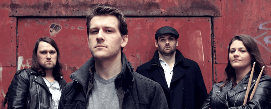 3 white men and one white woman standing in front of a rusty old wall. The first man has long hair and is wearing a leather jacket. The second is further forward in a grey coat. The third man is in a peaked cap and a black peacoat. The woman is in a leather jacket, holding drumsticks. They're all dressed for UK autumn weather, looking all mean and moody.